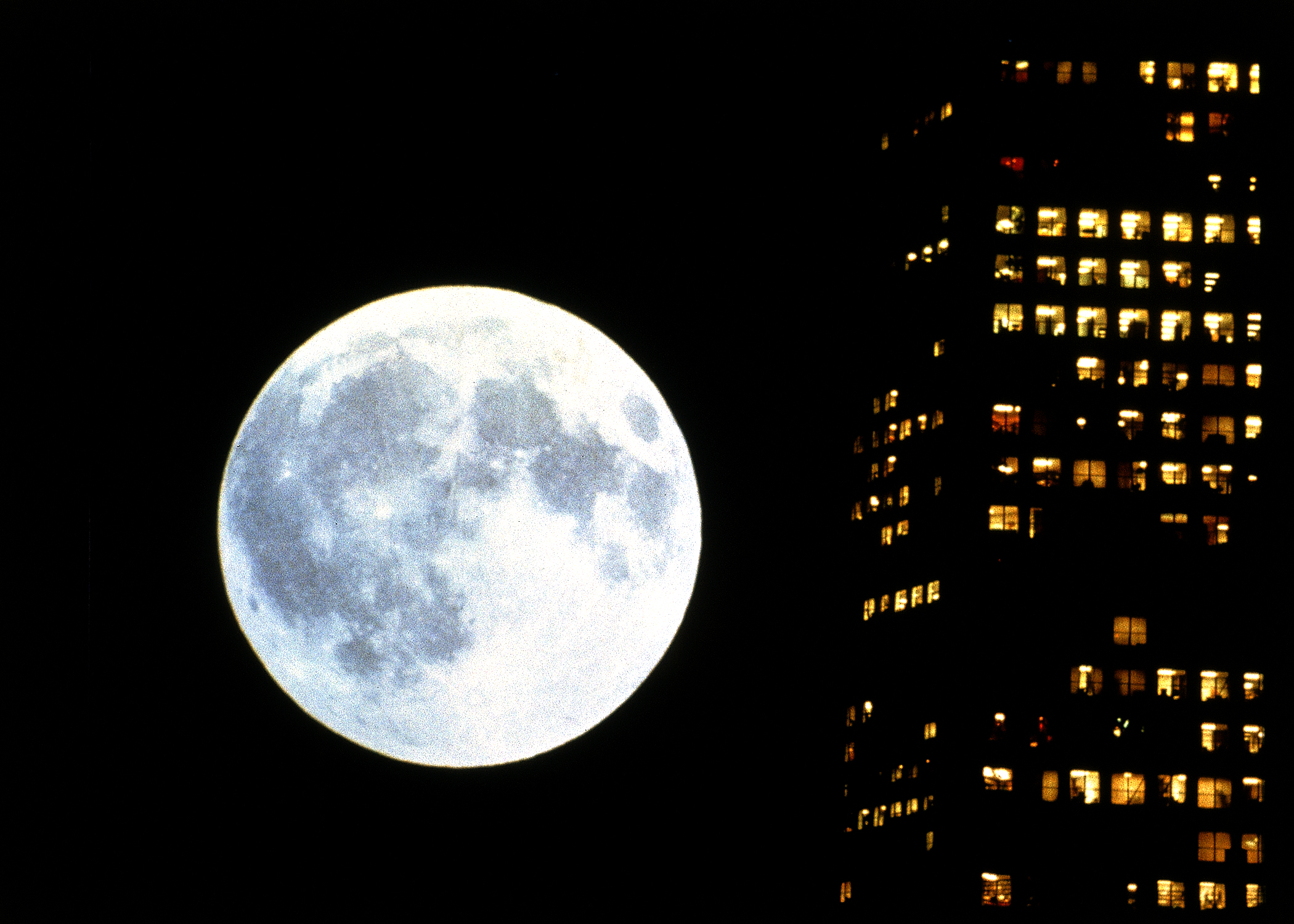 Kadr filmu “Koyaanisqatsi”, przedstawiający księżyc świecący w pełni po prawej stronie widać świecące okna wieżowca