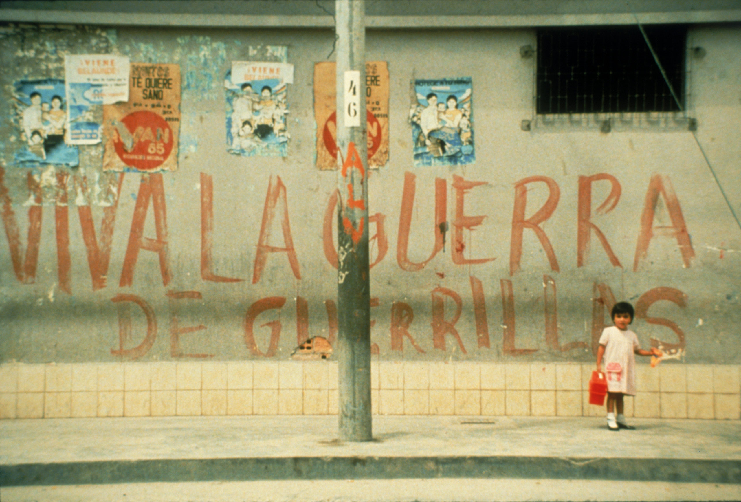 Kadr filmu "Powaqqatsi" przedstawiający małą dziewczynkę stojącą na chodniku, za nią jest szara ściana, a na niej namalowany czerwoną farbą napis