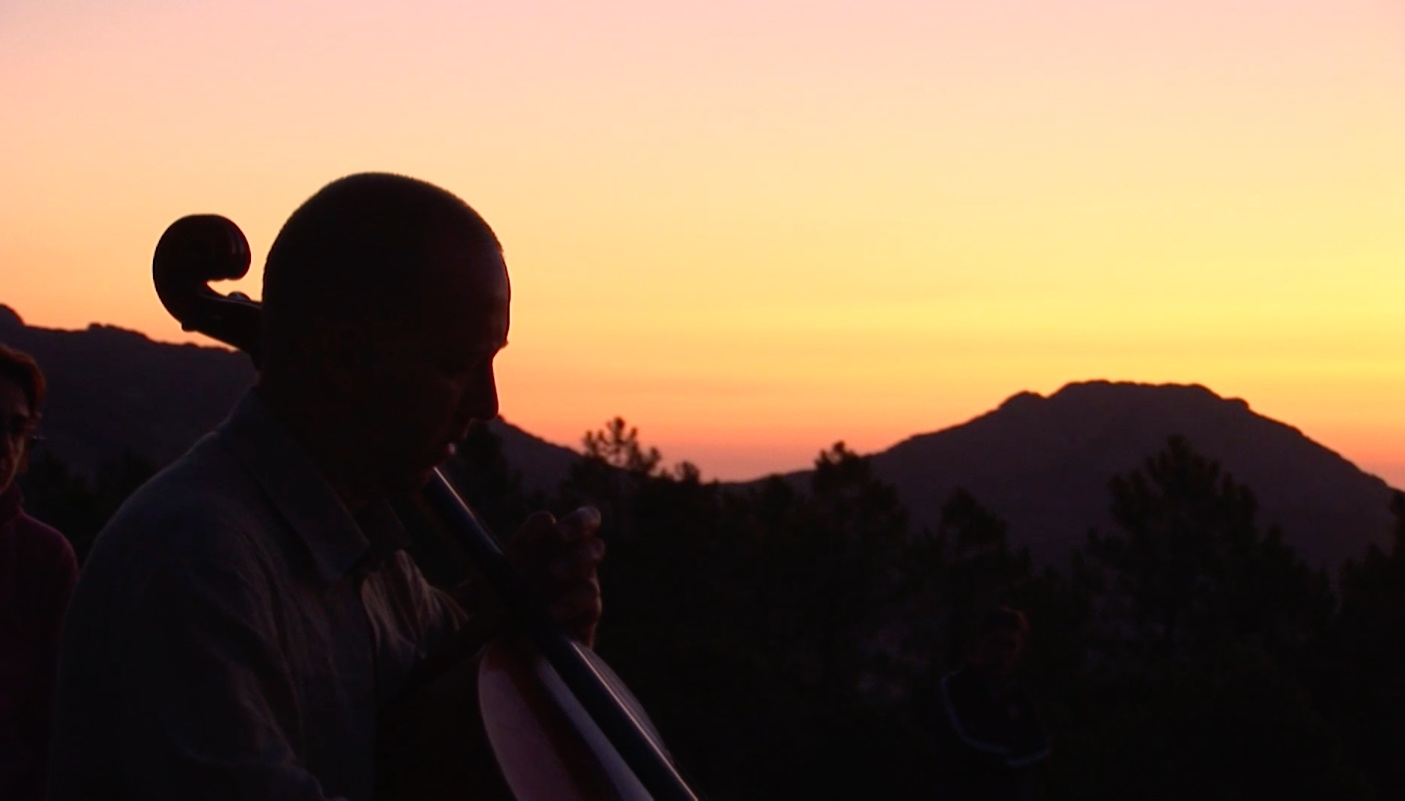Fragment filmu "Berchidda Live", przedstawiający ciemne kontury mężczyzny grającego na wiolonczeli, w tle widzimy obrys gór i zachód słońca.