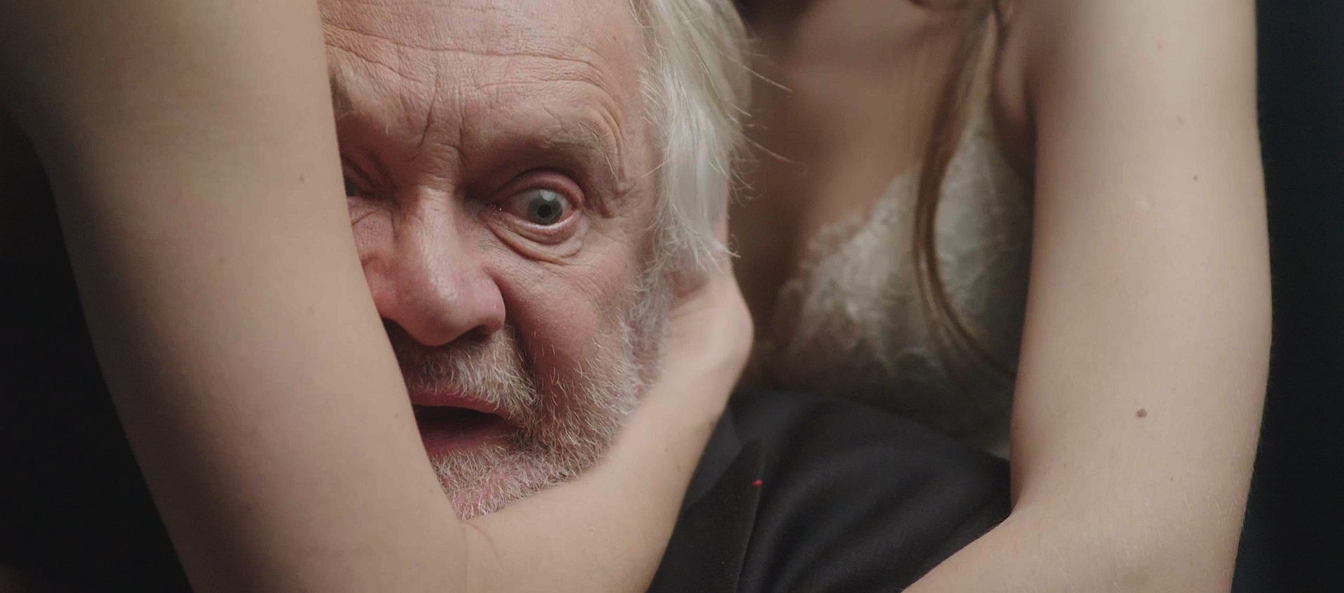 The frame of the film “I am a fictional character”, showing a close-up on the face of an elderly man who is hugged by women in their underwear, the protagonist looks shocked